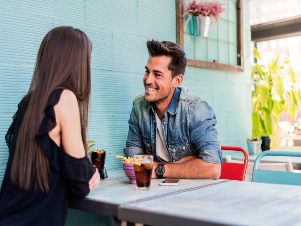 A picture of a couple sitting together while having drinks. The screen displays a website for finding Gottman Method Couples Therapists in New York City, with a search bar and a list of therapists' names and locations. The image conveys the idea of using technology to search for and find a therapist in the city, with the couple appearing happy and connected while doing so.