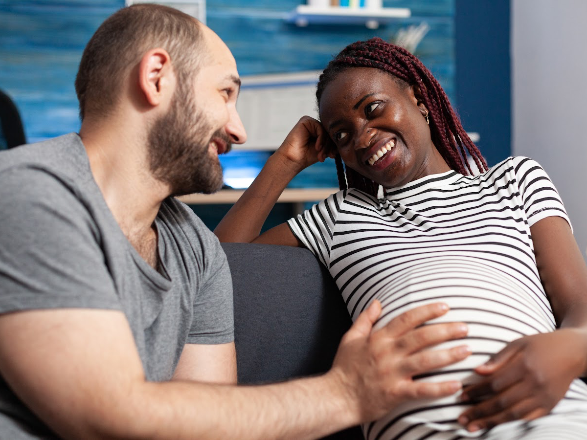 A happy interracial couple sits on the couch, engaging in open and honest communication. The pregnant wife shares her thoughts and feelings, while the attentive husband listens empathetically. Their successful marriage therapy at Loving at Your Best Marriage and Couples Counseling has helped them overcome the blame game and strengthen their relationship.