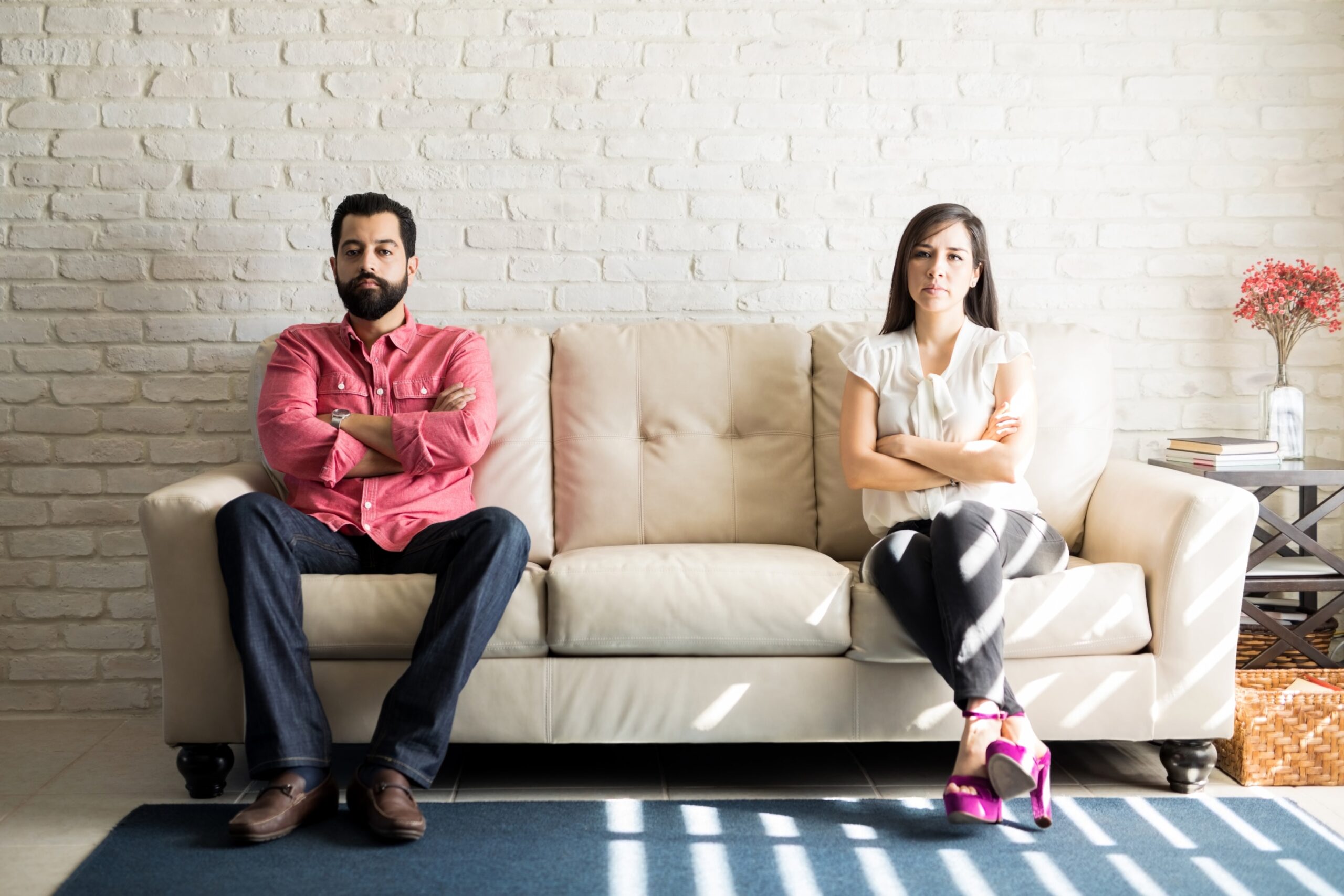 Couple sitting at opposite ends of couch looking conflicted despite apparently comfortable home, showing how affairs happen even in satisfying relationships.